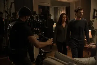 Camera operator with steadycam closely follows two actors performing a scene on a film set with crew and equipment visible, under warm, focused lighting.