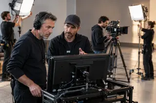 Chef opérateur concentré devant un moniteur sur un plateau de tournage avec le réalisateur, équipe technique à l’arrière-plan ajustant caméra et éclairage, ambiance professionnelle et chaleureuse.